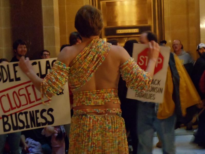 The front of this UW-Madison student's sign says Even I Look Less Ridiculous Than Scott Walker. He is wearing a grass skirt and toga-type top made entirely of candy necklaces. The back of the sign: Middle Class: CLOSED FOR BUSINESS